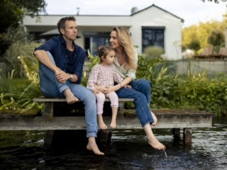 Junge Familie mit einem Mädchen sitzt vor dem Haus am See und lässt die Füsse baumeln